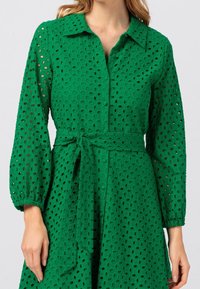 Grön klänning i ögla tyg med krage, långa puffärmar, bälte i midjan och utsvängd kjol.