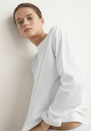 Young woman leaning against a wall wearing a white long-sleeve shirt and light-colored pants, looking into the camera with a neutral expression.