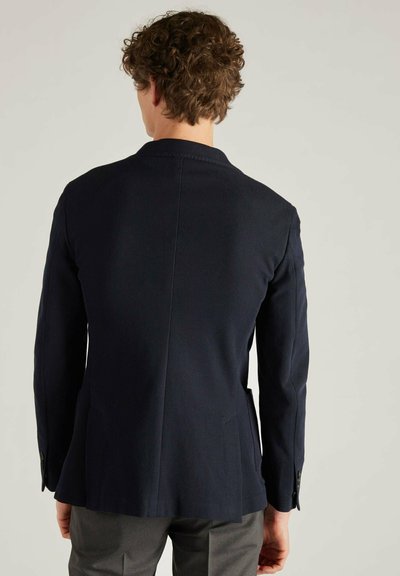 Navy Blazer aus strukturiertem Stoff, schlank geschnitten, mit einem Schlitzt hinten und schwarzen Knopf-Details, kombiniert mit grauen Hosen.