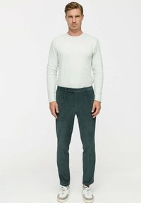 Egy férfi áll, szemben a kamera, fehér hosszú ujjú inget, sötétzöld kordbársony nadrágot és fehér sportcipőt visel, egy sima háttér előtt.