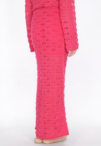 Frau trägt einen langen, leuchtend pinken gehäkelten Rock und ein passendes Oberteil, steht mit einer sichtbaren Hand und kombiniert dazu gewebte Plateauschuhe.