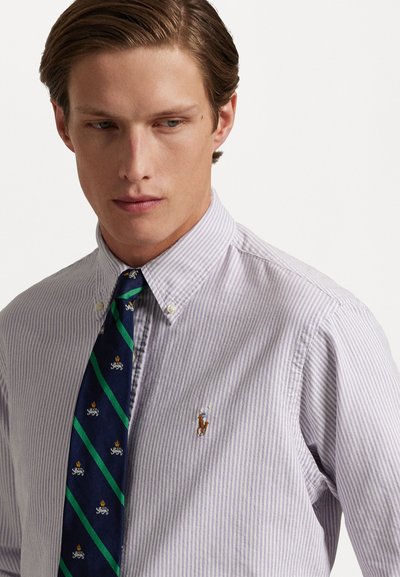 Chemise rayée violette claire et blanche avec un col boutonné, dotée d'un petit logo de cheval. Associée à une cravate marine, accentuée de rayures et de motifs verts.
