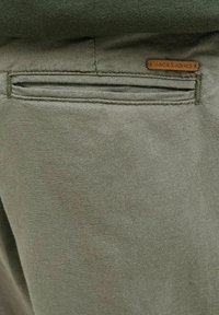 Olive zelene bombažne hlače imajo zadnji žep s subtilnim detajlom oznake, kar prikazuje gladko, teksturirano tkanino in poenostavljen dizajn.