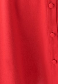 Röd tyg med en mjuk yta, som har en rad av små, runda knappar längs ena sidan, vilket ger ett strukturerat designelement.