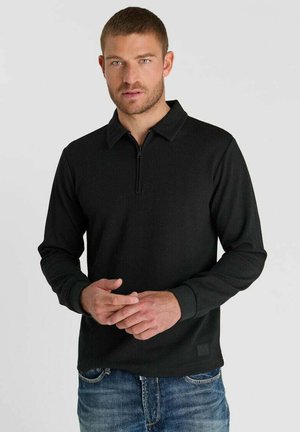 Uomo con capelli corti che indossa una maglia nera a maniche lunghe con zip e jeans blu, in piedi davanti a uno sfondo chiaro e semplice.