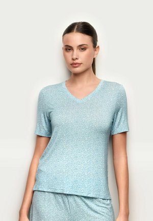 Junge Frau mit zurückgekämmtem Haar, die ein hellblaues, kurzärmeliges V-Ausschnitt-Oberteil und passende Hosen mit dezentem Leopardenmuster trägt.