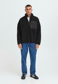 Sudadera de forro polar negra con media cremallera, que presenta una superficie texturizada similar a la de un pelaje, y un bolsillo en el pecho en negro de contraste. Combinada con unos vaqueros azules.