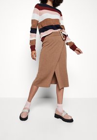 Žena oblečená ve pruhovaném svetru, široké camel sukni, růžových ponožkách a robustních mokasínách s pastelovými popruhy, stojící na bílém pozadí.