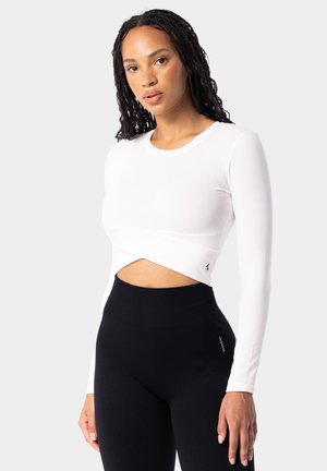Bílý dlouhý rukáv cropped top s kulatým výstřihem a zkrouceným detailem vpředu, kombinovaný s černými legínami s vysokým pasem, které mají nenápadné logo.