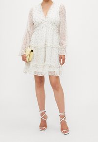 Witte bloemenjurk met lange doorschijnende mouwen, ruches langs de lagen en een diepe V-hals. Gecombineerd met witte sandalen met bandjes en een gele clutch.