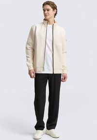 Crèmekleurig bomberjack met gestructureerde stof, zwarte rits en geribbelde manchetten, gecombineerd met een wit T-shirt, zwarte broek en lichte sneakers.