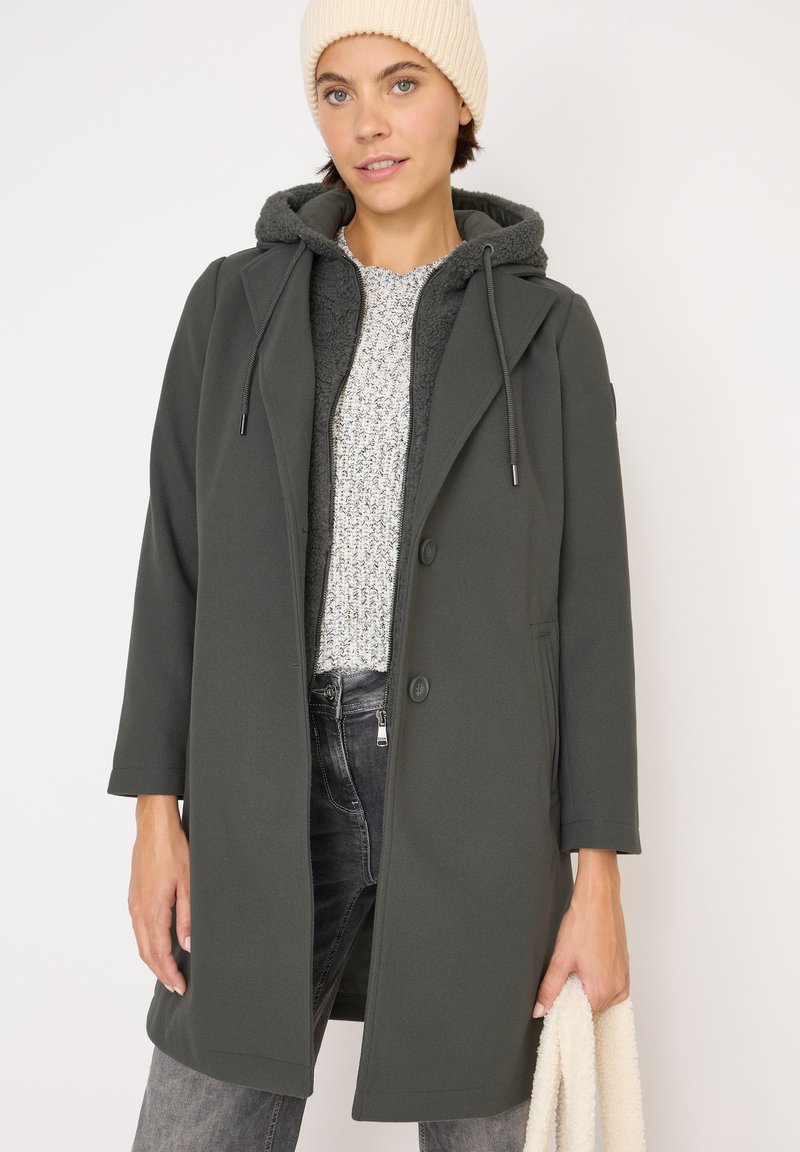 Cappotto verde scuro con cappuccio, dotato di una morbida texture interna, due bottoni e tasche laterali. Abbinato a un maglione lavorato a maglia e jeans.