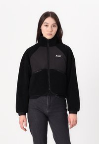 Giacca nera corta con texture in pile sul corpo e tessuto liscio sulle spalle, dotata di chiusura con zip e dettaglio del logo.