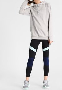 Kvinna iklädd en ljusgrå hoodie, svarta leggings med blå och ljusblå geometriska mönster, samt grå träningsskor, som står framför en enhetlig bakgrund.
