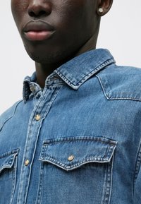 Chemise en denim bleu avec un col structuré, deux poches poitrine avec fermeture à bouton et une texture légèrement délavée.
