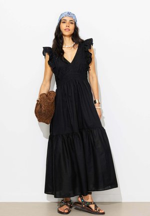 Femme aux longs cheveux foncés portant une robe maxi noire à volants, un foulard bleu à fleurs, un collier de coquillages, tenant un sac à motif léopard et portant des sandales cloutées.
