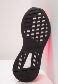 Svart gummiskosula med texturerat rutmönster, med en vit mellansula och präglad logotyp. Positionerad mot en ljus bakgrund.