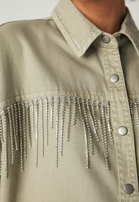 Kaki marškiniai su sidabriniais deimantinio lurekso pakraščiais aplink krūtinę, turintys apykaklę ir metalinius spaudimo mygtukus.