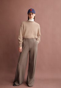 Beige, flauschiger Pullover mit Rippenbündchen kombiniert mit hochgeschnittenen, weit geschnittenen taupefarbenen Hosen. Eine marineblaue Mütze mit roten Akzenten vervollständigt den Look.