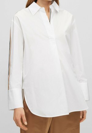 Woman wearing a white long-sleeve blouse with side slits and tan, black, and white striped detailing on the sleeves, paired with brown pants.