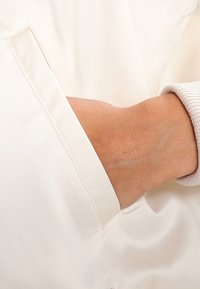 Cream-colored fabric with a smooth texture features a hand reaching into a neatly stitched side pocket. The sleeve has a ribbed cuff.