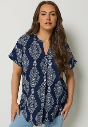 Kobieta o długich brązowych włosach, ubrana w granatową bluzkę z krótkim rękawem z białym, misternym wzorem, w połączeniu z jasnoniebieskimi dżinsami, stojąca na tle gładkiej ściany.