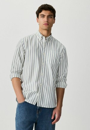 Mladý muž s tmavými kučeravými vlasmi, oblečený v bielej košeli s dlhým rukávom so svetlomodrými zvislými pruhmi a modré džínsy, stojaci pred jednoduchým pozadím.