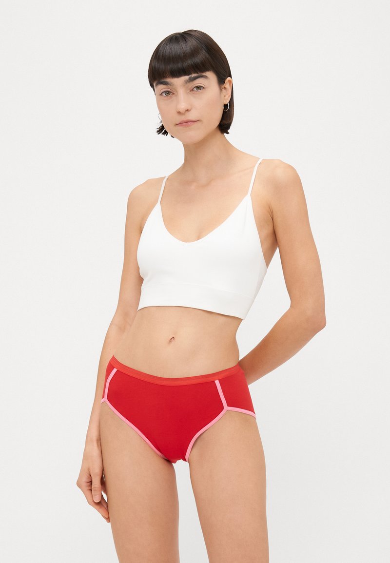 Fehér bralette vékony pántokkal, piros alsóneművel párosítva, pink kiegészítésekkel és varrással. Simaság és testhezálló kialakítás jellemzi.