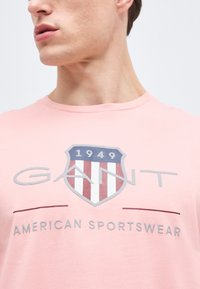 Κοντινό πλάνο ατόμου που φοράει ροζ πουκάμισο με το λογότυπο GANT, έμβλημα ασπίδας με το "1949" και κείμενο "AMERICAN SPORTSWEAR" στο στήθος.