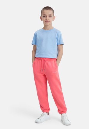 Dreng står med hænderne i lommerne iført lys blå t-shirt, koral sweatpants og hvide sneakers mod en hvid baggrund.