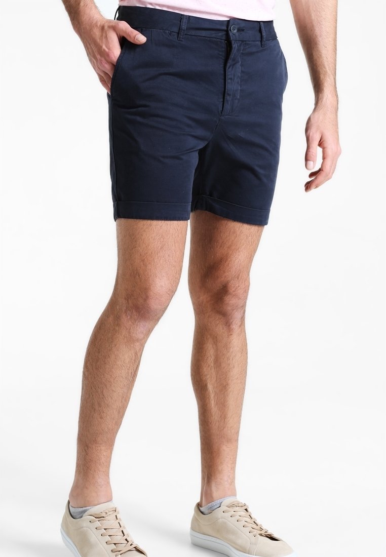 Shorts en coton bleu marine foncé avec une fermeture à bouton et fermeture éclair à l'avant, présentant des ourlets roulés et des poches latérales. Portés avec des chaussures beige clair.