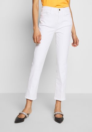 Persoon draagt witte jeans met rechte pijpen, oranje top en zwarte puntige ballerina's met strassriempjes, staand tegen een lichte achtergrond.