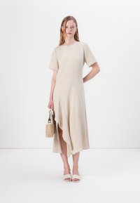 Beige, knelang kjole med korte ærmer, med asymmetrisk kant og tekstureret stof. Modellen holder en lille vævet taske og har blege sandaler på.
