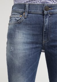 Denim jeans in a medium blue wash with subtle fading, a button closure and multiple pockets, featuring distressed detailing near the thigh.