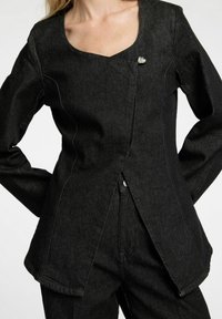 Svart denimjacka med fyrkantig halsringning, långa ärmar, framknäppning och en silverfärgad hjärtformad detalj på bröstet. Strukturerad tyg.