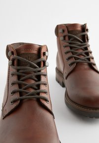 Hnedé kožené členkové čižmy s textúrovaným povrchom, kovovými očkami a tmavohnedými šnúrkami. Majú pevný podrážku a kontrastný detail na golieri.