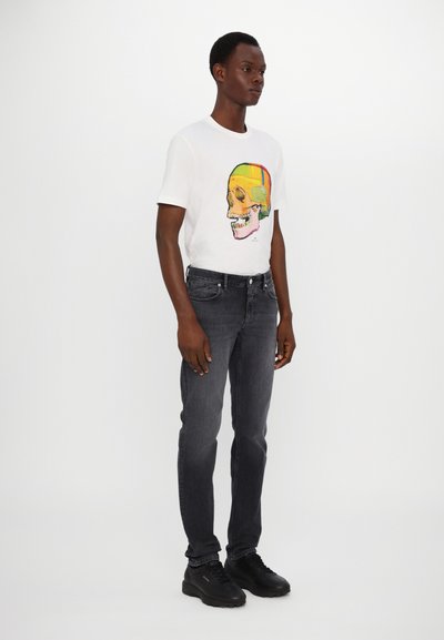 Camiseta blanca con un estampado colorido de calavera en el centro, combinada con jeans gris oscuro y zapatillas negras. Diseño sencillo, ajuste casual.