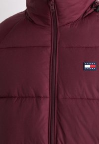 Giubbino imbottito color bordeaux con texture trapuntata e colletto alto. Presenta una zip frontale e una piccola toppa con il logo 'Tommy Jeans'.