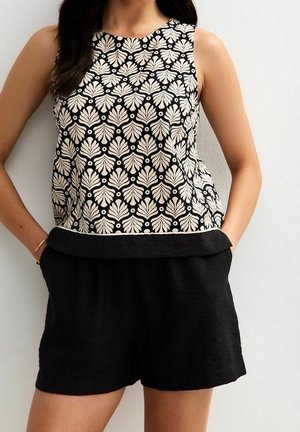 Top sin mangas con patrón en negro y beige combinado con pantalones cortos negros lisos, manos casualmente en los bolsillos contra un fondo blanco liso.