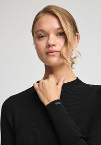 Svart ribbad tröja med långa ärmar, hög halsringning och en texturerad yta, med en liten svart etikett på ärmen.