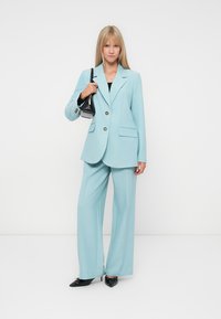 Light blue suit featuring a tailored blazer and wide-leg pants. Textured fabric, two front pockets, and gold button details. Black handbag.