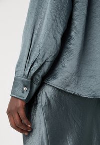Main portant une chemise à manches longues avec poignets boutonnés en tissu froissé bleu canard, avec une jupe froissée assortie visible sous l'ourlet de la chemise.