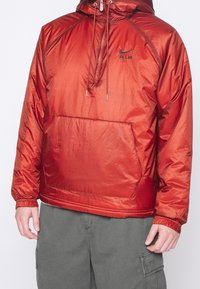 Röd nylonpulloverjacka med huva, utrustad med justerbar dragsko, en framficka och en svart broderad logotyp på bröstet.