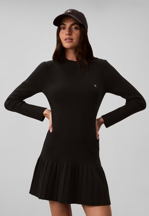 Donna che indossa un vestito nero a costine a maniche lunghe con orlo plissettato e un berretto nero, in piedi con le mani sui fianchi su sfondo grigio.