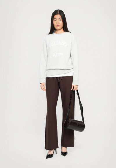 Femme portant un sweatshirt gris clair "Voltaire Paris", un pantalon évasé marron foncé, des escarpins noirs à bout pointu, tenant un sac cylindrique noir.