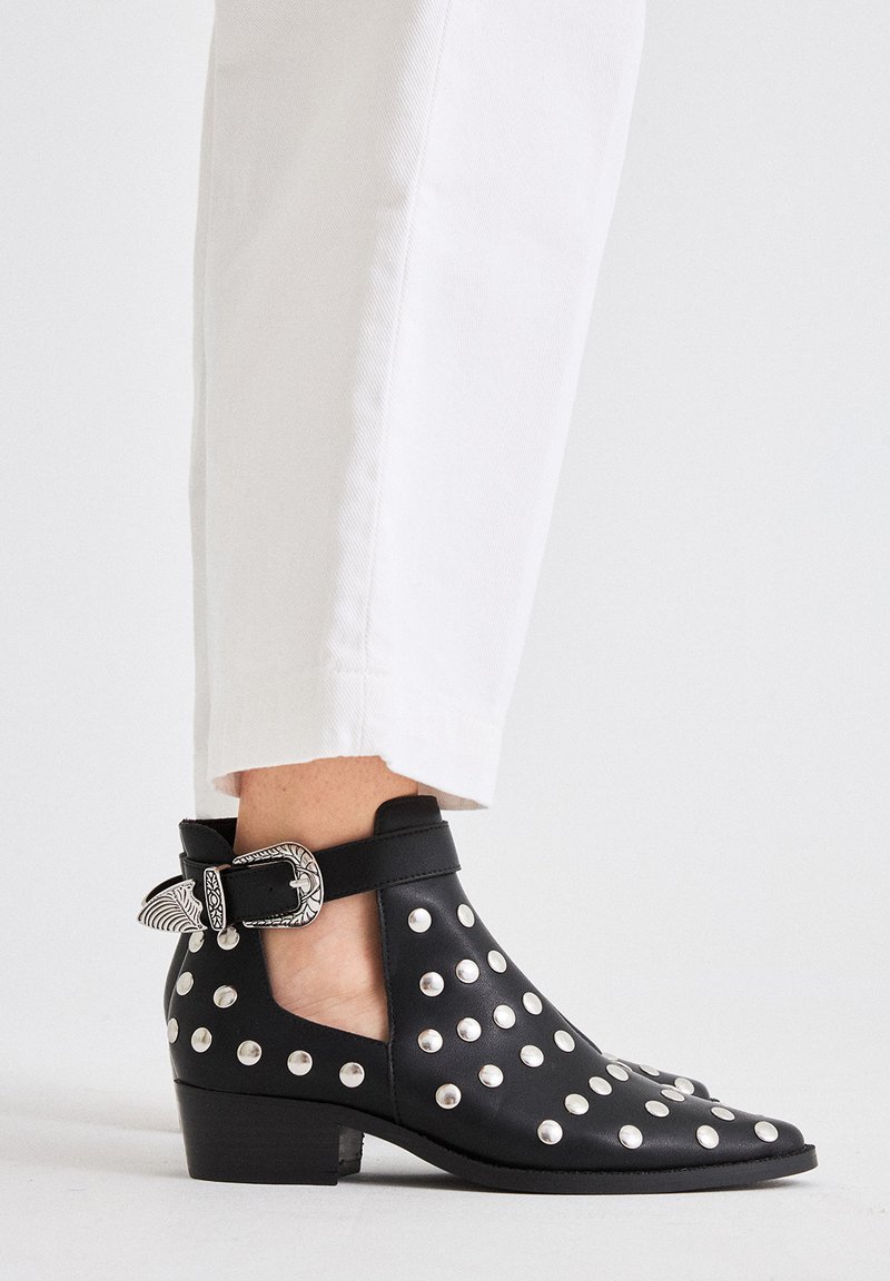 Botín negro de cuero con adornos de tachuelas plateadas, tacón bajo y hebilla decorativa en el lateral. Combinado con pantalones blancos con puños.