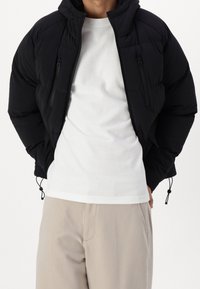 Giacca piumino nera con cappuccio, dotata di due tasche con zip e cordini regolabili. Indossata sopra una camicia bianca e pantaloni beige chiaro.