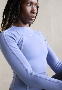 Mlada žena s pletenicom, koja nosi svijetloplavu dugorukavu Nike majicu, izgleda koncentrirano i blago je okrenuta sa strane u neutralnom okruženju.