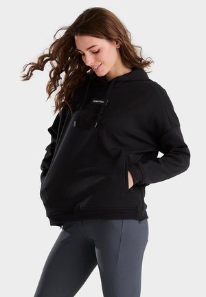 Mujer con cabello largo y castaño que lleva una sudadera negra con capucha y las manos en los bolsillos, y leggings gris oscuro, mirando hacia abajo y sonriendo.