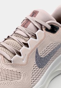 Roza atletska cipela s mrežastim materijalom, teksturiranom površinom i srebrnim sjajnim Nike logom. Ima vezice i uzorčani jezik.